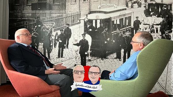Kübi und Kurt sitzen vor einem historischen Foto im Betriebshof Heinrich-Alfes-Straße